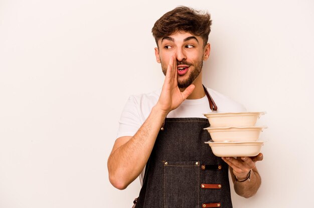 Jonge Spaanse klerk man met een tupperware geïsoleerd op een witte achtergrond zegt een geheim heet remnieuws en kijkt opzij