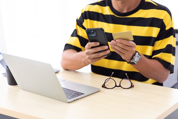 Jonge spaanse man die creditcard gebruikt om online te betalen met smartphone en laptopcomputer thuis of op creatieve werkplek.
