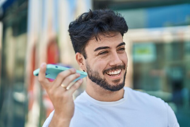 Jonge spaanse man die lacht zelfverzekerd luisterend audiobericht door de smartphone op straat