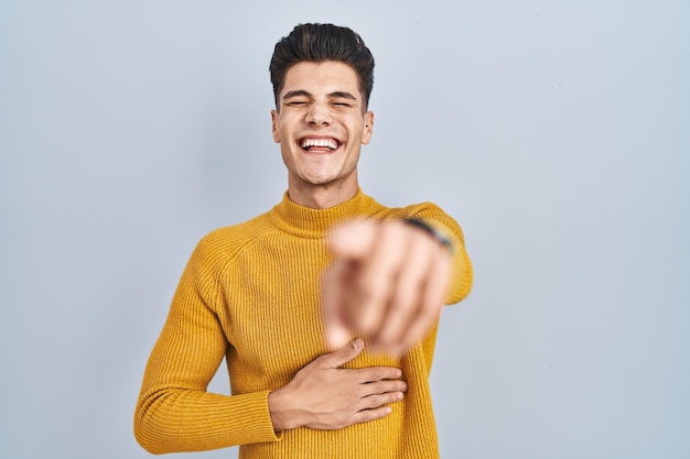 Jonge spaanse man die over een blauwe achtergrond staat en je uitlacht en met de vinger naar de camera wijst met de hand over de schaamte van het lichaam