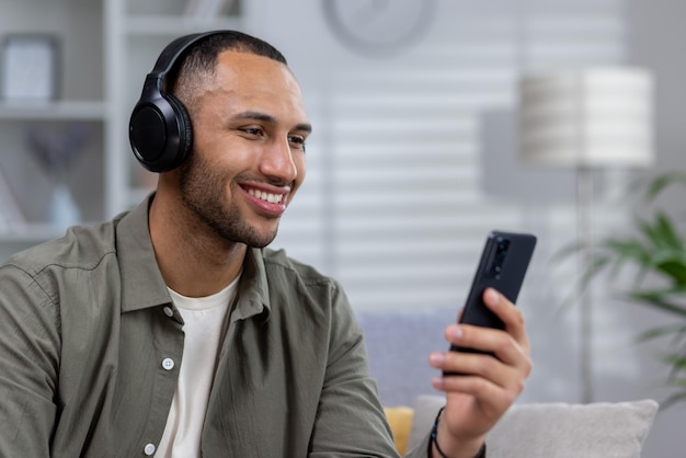 Jonge spaanse man die thuis zit met een koptelefoon die de telefoon in de hand houdt en naar muziek luistert