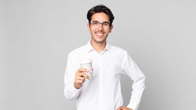 Jonge Spaanse man die vrolijk lacht met een hand op de heup en zelfverzekerd en een afhaalkoffie vasthoudt