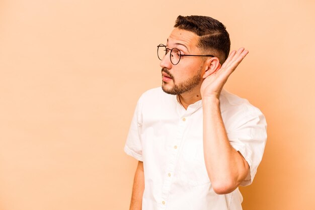 Jonge Spaanse man geïsoleerd op beige achtergrond probeert te luisteren naar een roddel