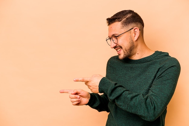 Jonge Spaanse man geïsoleerd op beige achtergrond wijst met duim vinger weg lachend en zorgeloos