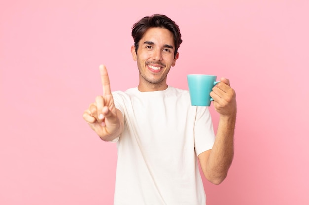 Jonge Spaanse man lacht en ziet er vriendelijk uit en toont nummer één en houdt een koffiemok vast