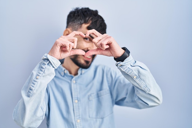 Jonge spaanse man met baard die over blauwe achtergrond staat en hartvorm doet met hand en vingers glimlachend kijkend door teken