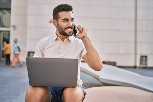 Jonge Spaanse man met behulp van laptop praten op de smartphone op straat