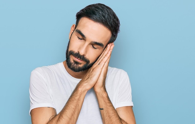 Jonge spaanse man met casual wit t-shirt slapend moe dromend en poserend met de handen in elkaar terwijl hij glimlacht met gesloten ogen.