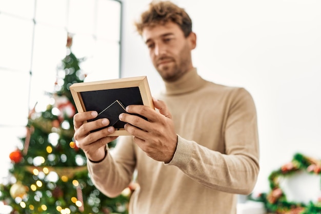 Jonge spaanse man met foto die thuis bij de kerstboom staat