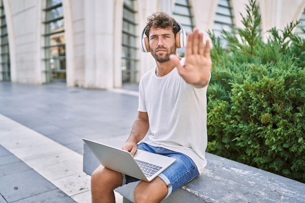 Jonge spaanse man met koptelefoon en laptop op straat met open hand stopbord doen met serieus en zelfverzekerd uitdrukkingsverdedigingsgebaar