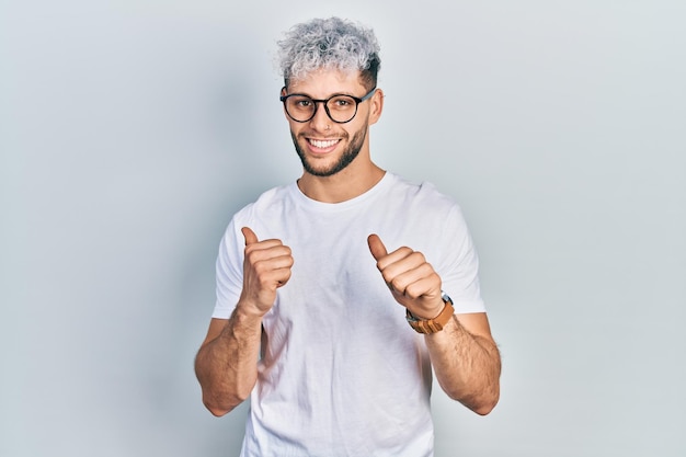 Jonge spaanse man met modern geverfd haar in wit t-shirt en bril naar achteren wijzend met hand en duimen omhoog glimlachend zelfverzekerd