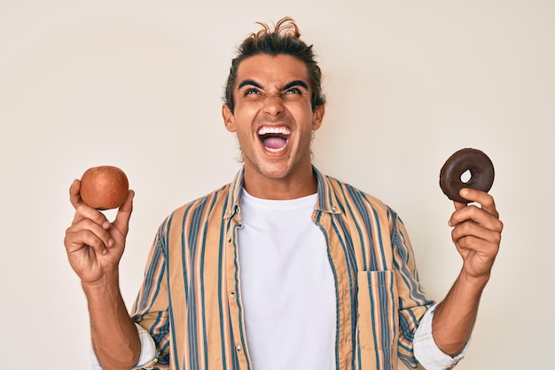 Foto jonge spaanse man met rode appel en donut boos en boos schreeuwend gefrustreerd en woedend schreeuwend van woede die omhoog kijkt