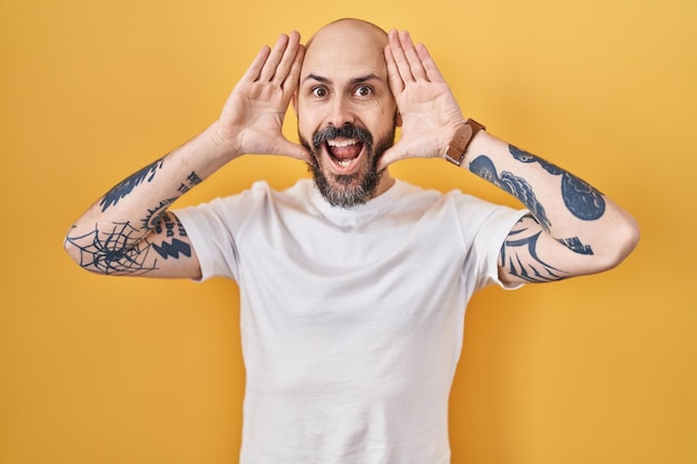 Jonge spaanse man met tatoeages die over een gele achtergrond staan glimlachend vrolijk spelen peek a boo met handen die zijn gezicht verrast en opgewonden laten zien
