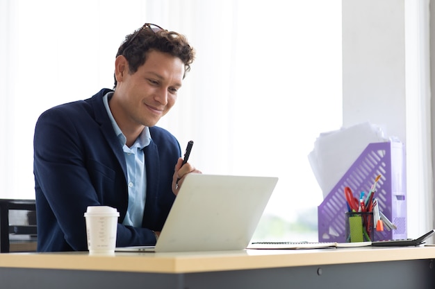 Jonge Spaanse man zit koffie te drinken en werkt op laptopcomputer op creatieve kantoor aan huis.