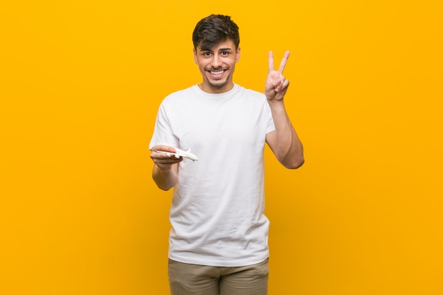 Jonge Spaanse mens die een vliegtuigpictogram houdt dat overwinningsteken toont en breed glimlacht.