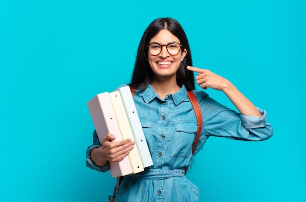 Jonge Spaanse studentenvrouw die zelfverzekerd glimlachen wijst naar eigen brede glimlach, positieve, ontspannen, tevreden houding