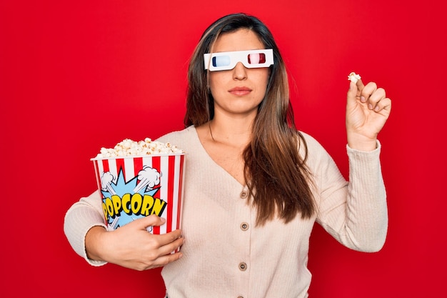 Jonge Spaanse vrouw die 3D-filmbril draagt en popcorn eet op rode achtergrond met een zelfverzekerde uitdrukking op een slim gezicht die serieus denkt