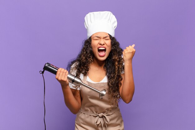 Jonge spaanse vrouw die agressief schreeuwt met een boze uitdrukking of met gebalde vuisten om succes te vieren. chef-kok concept