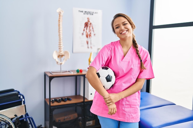 Jonge spaanse vrouw die bij de fysiotherapiekliniek werkt en voetbalbal vasthoudt die er positief en gelukkig uitziet en glimlacht met een zelfverzekerde glimlach die tanden laat zien