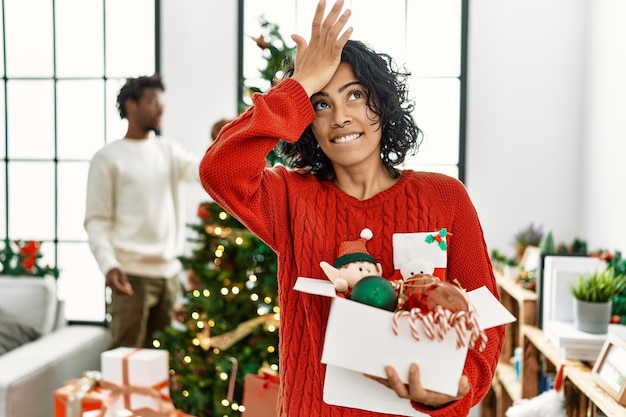 Jonge spaanse vrouw die bij de kerstboom staat met versiering verrast met de hand op het hoofd voor een fout, onthoud fout. vergeten, slecht geheugenconcept.