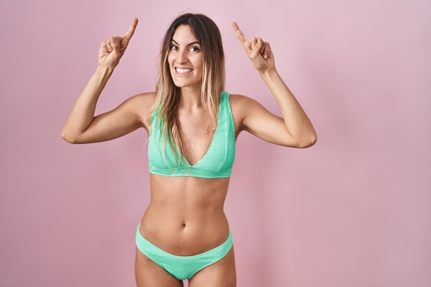 Jonge spaanse vrouw die bikini draagt over roze achtergrond glimlachend verbaasd en verrast en omhoog wijzend met vingers en opgeheven armen.