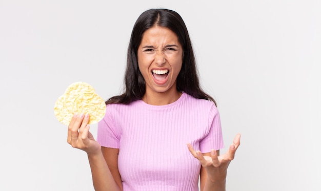 Jonge spaanse vrouw die boos, geïrriteerd en gefrustreerd kijkt en een rijstkoekje vasthoudt. dieet concept