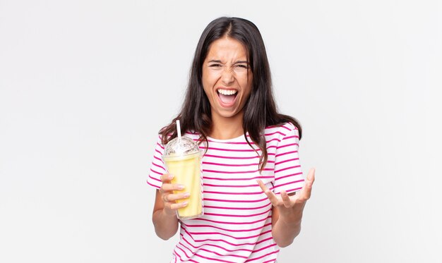 Jonge Spaanse vrouw die boos, geïrriteerd en gefrustreerd kijkt en een vanila smoothy milkshake vasthoudt