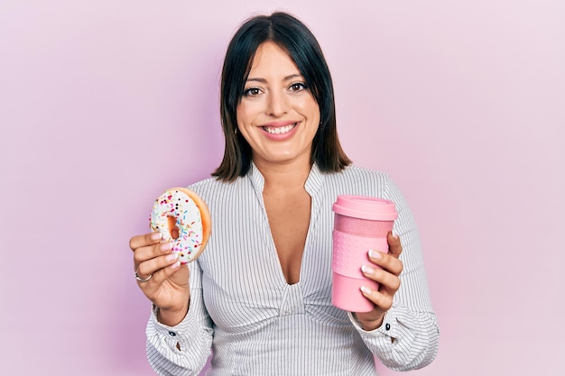 Jonge spaanse vrouw die donut eet en koffie drinkt glimlachend met een gelukkige en koele glimlach op het gezicht met tanden