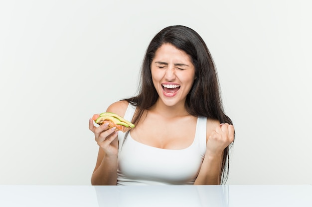 Jonge Spaanse vrouw die een avocadotoost houden onbezorgd en opgewekt toejuichen. Overwinning concept.