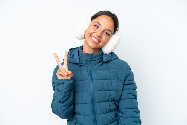 Jonge spaanse vrouw die een de winteroorbeschermers draagt die op witte achtergrond wordt geïsoleerd die en overwinningsteken glimlacht toont