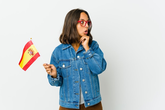 Jonge spaanse vrouw die een vlag houdt die op witte achtergrond wordt geïsoleerd die voor geluk bidt, verbaasd en mond opent die naar voorzijde kijkt.