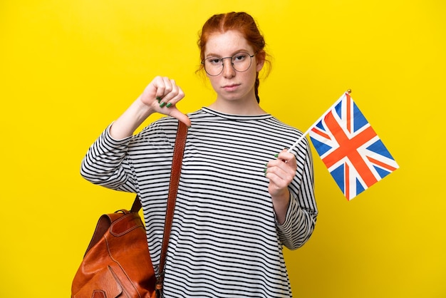 Jonge spaanse vrouw die een vlag van het Verenigd Koninkrijk houdt die op gele achtergrond wordt geïsoleerd die duim neer met negatieve uitdrukking toont