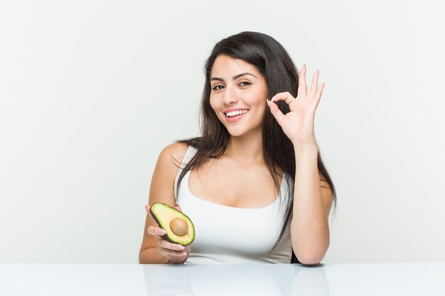 Jonge Spaanse vrouw die een vrolijk en zeker avocado houdt die ok gebaar toont.