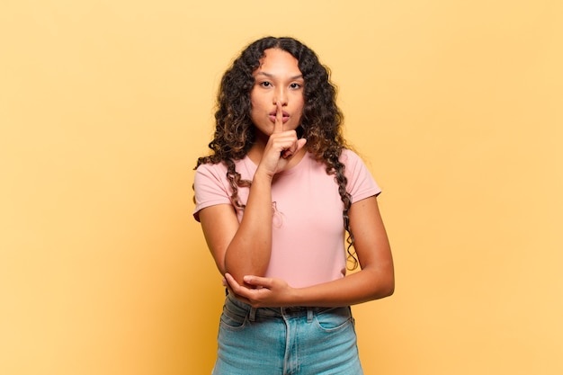 Jonge Spaanse vrouw die er serieus uitziet en kruist met de vinger tegen de lippen gedrukt die stilte of stilte eist en een geheim bewaart