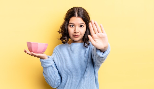 Jonge spaanse vrouw die er serieus uitziet en open palm toont die een stopgebaar maakt. lege kom concept