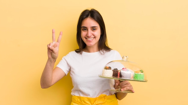 Foto jonge spaanse vrouw die lacht en er vriendelijk uitziet met het concept van nummer twee zelfgemaakte taarten