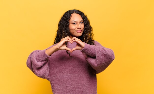 Jonge Spaanse vrouw die lacht en zich gelukkig, schattig, romantisch en verliefd voelt, maakt hartvorm met beide handen