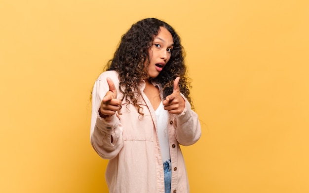 Jonge Spaanse vrouw die lacht met een positieve, succesvolle, gelukkige houding die naar de camera wijst en een pistool met handen maakt