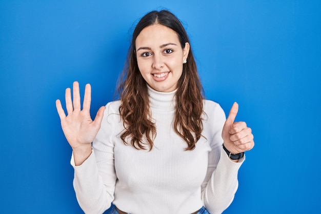 Jonge spaanse vrouw die over een blauwe achtergrond staat en met vingers nummer zes naar boven wijst terwijl ze zelfverzekerd en gelukkig glimlacht.