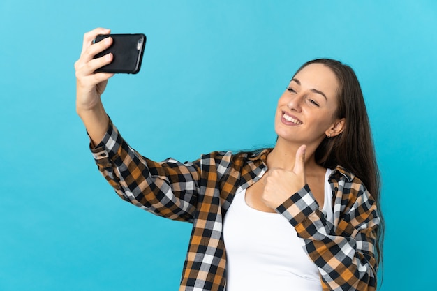 Jonge Spaanse vrouw die over geïsoleerde blauwe muur een selfie met mobiele telefoon maakt