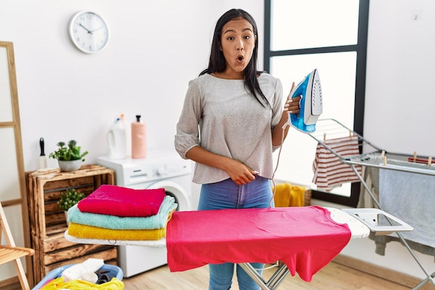 Jonge spaanse vrouw die thuis kleren strijkt, bang en verbaasd met open mond voor een verbaasd ongeloofgezicht