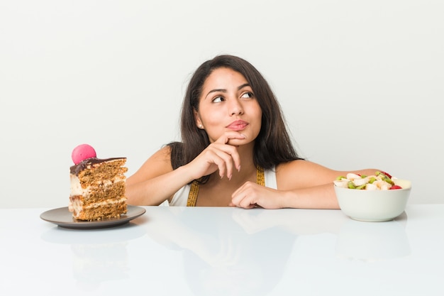 Jonge Spaanse vrouw die tussen cake of fruit kiest die zijdelings met twijfelachtige en sceptische uitdrukking kijkt.