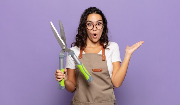 Jonge Spaanse vrouw die verbaasd en geschokt kijkt, met open mond een voorwerp vasthoudt met een open hand op de zijkant