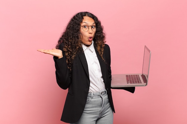 Jonge spaanse vrouw die verrast en geschokt kijkt, met open mond terwijl ze een object vasthoudt met een open hand aan de zijkant. laptopconcept