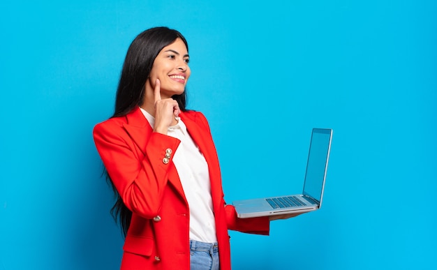 Jonge spaanse vrouw die vrolijk lacht en dagdroomt of twijfelt, opzij kijkend. laptopconcept