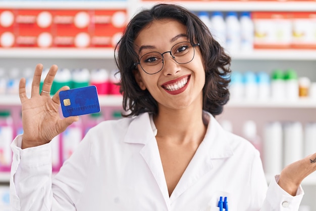 Jonge spaanse vrouw die werkt bij apotheekdrogisterij met creditcard die prestatie viert met een gelukkige glimlach en winnaarsuitdrukking met opgeheven hand