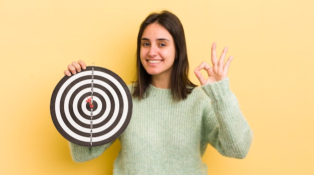 Jonge Spaanse vrouw die zich gelukkig voelt en goedkeuring toont met een goed gebaar darts doelconcept