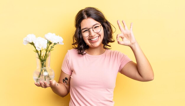 Jonge spaanse vrouw die zich gelukkig voelt, goedkeuring toont met een goed gebaar. bloemen pot concept