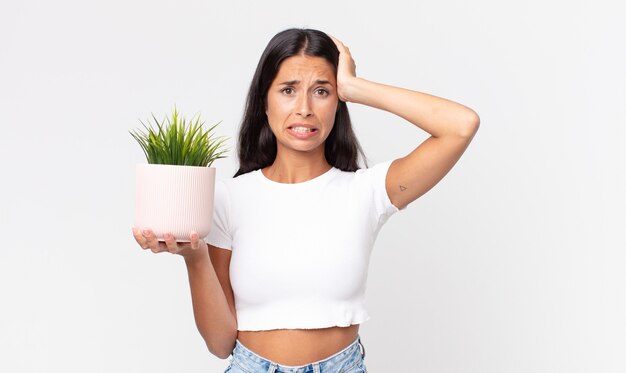 Jonge Spaanse vrouw die zich gestrest, angstig of bang voelt, met de handen op het hoofd en een decoratieve kamerplant vasthoudt