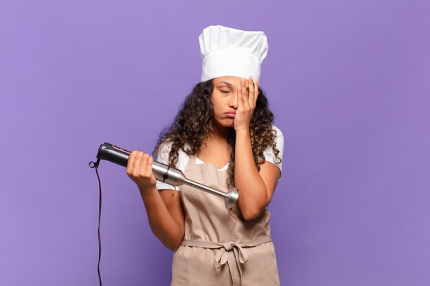 Jonge Spaanse vrouw die zich verveeld, gefrustreerd en slaperig voelt na een vermoeiende, saaie en vervelende taak, gezicht met de hand vasthoudend. chef-kok concept